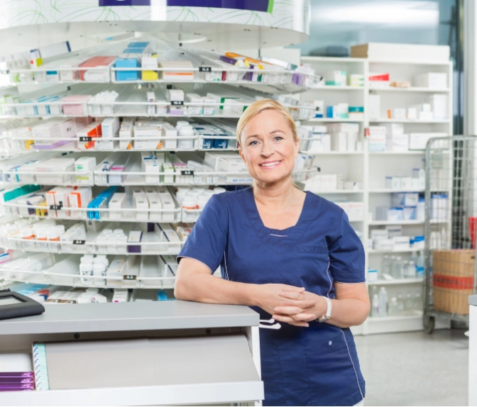 Smiling pharmacist in blue scrubs stands at a counter in a pharmacy with shelves of medicine behind her.