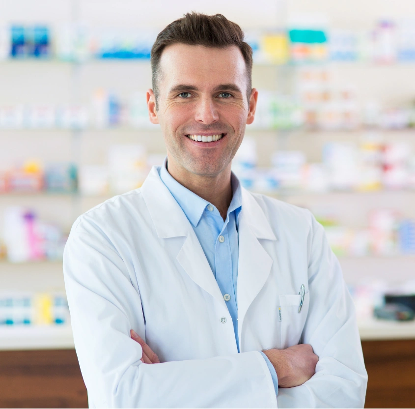 Smiling pharmacist in a white coat stands with arms crossed in a pharmacy.