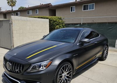 A black luxury sports car with yellow accents, complete with sleek window tinting, is parked in a driveway near residential buildings and palm trees.